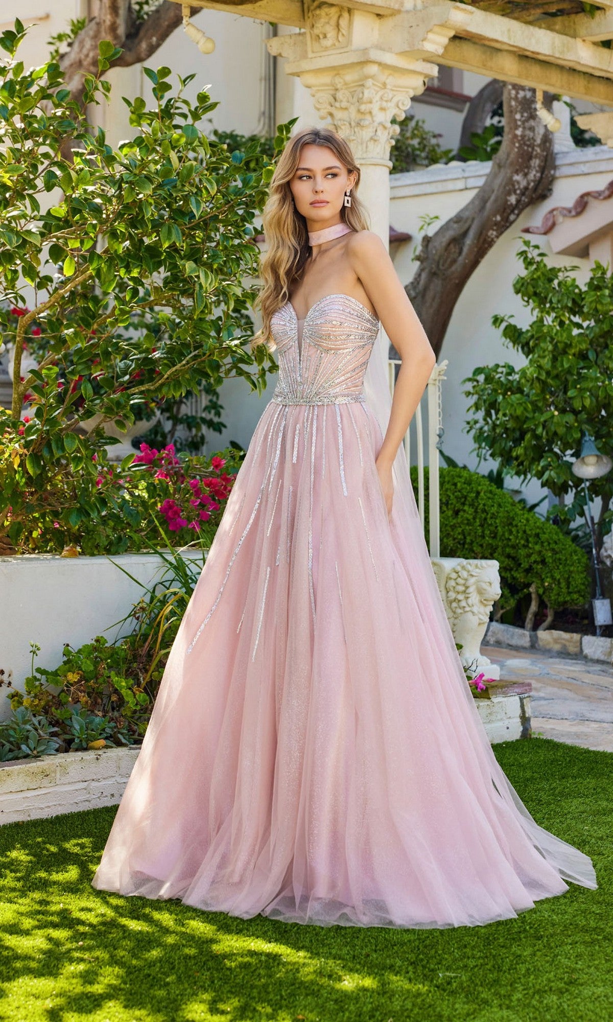 A woman wearing the Juliet JT2620U long prom dress—strapless, light pink tulle with a fitted, rhinestone-embellished bodice—stands outdoors on green grass near lush plants and a stone structure.